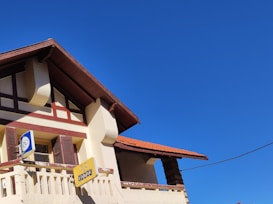 A traditional house with a sloped red-tiled roof and beige walls, featuring wooden shutters on the windows. Two signs are visible, one blue and one yellow, with symbols and text. The sky above is clear and deep blue.