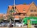 A rustic wooden building with a steep, orange-tiled roof stands prominently, housing a pharmacy and a hotel. Several flags hang from a pole near the building. In the foreground, parked cars line the street in front of a large green truck. The pharmacy is indicated by a green cross sign, and there are trees with green foliage scattered throughout the scene.