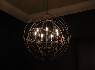 A spherical chandelier with multiple exposed light bulbs hangs from the ceiling. The fixture features a metal cage-like design with a modern, industrial aesthetic. The surrounding area is dimly lit, highlighting the warm glow emitted from the bulbs against a dark background.