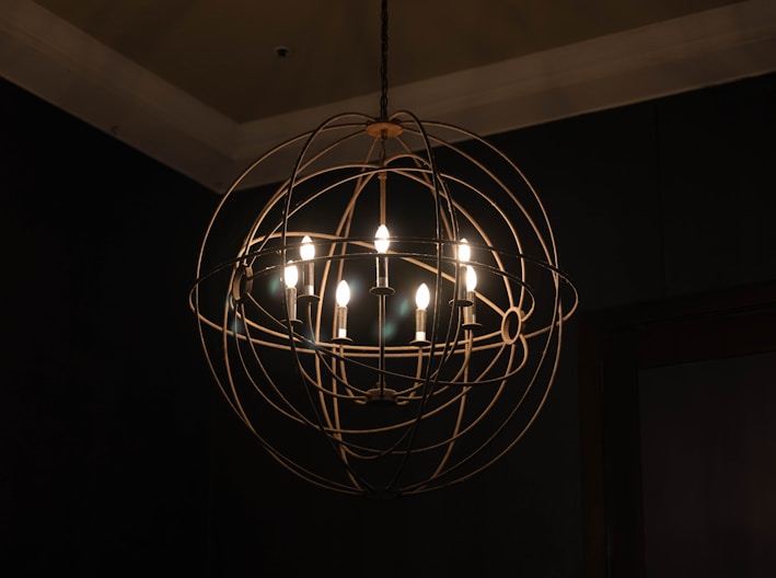 A spherical chandelier with multiple exposed light bulbs hangs from the ceiling. The fixture features a metal cage-like design with a modern, industrial aesthetic. The surrounding area is dimly lit, highlighting the warm glow emitted from the bulbs against a dark background.