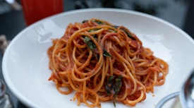 A plate of spaghetti topped with a rich red tomato sauce and garnished with fresh green basil leaves. The dish is served in a clean white bowl, and the focus is on the vibrant colors and textures of the pasta and sauce.