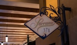 A decorative signage with the word 'Serrano' followed by the number '378'. The sign features elegant, swirling wrought iron details and is mounted on a textured wall. The background includes a ceiling with exposed wooden beams and hanging lights, contributing to a warm and inviting ambiance.