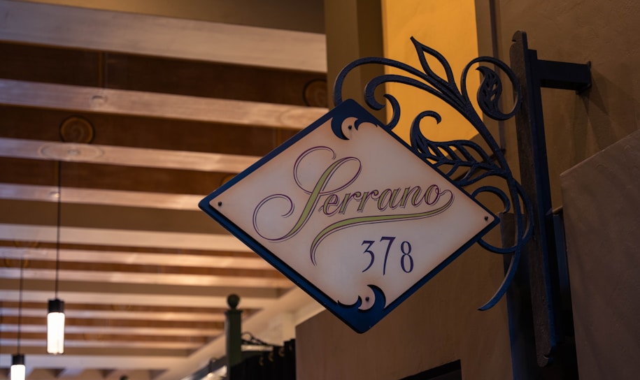 A decorative signage with the word 'Serrano' followed by the number '378'. The sign features elegant, swirling wrought iron details and is mounted on a textured wall. The background includes a ceiling with exposed wooden beams and hanging lights, contributing to a warm and inviting ambiance.