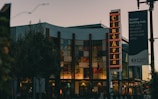 A theater building with a prominent vertical neon sign displaying 'Ciné Arts' situated at the front. The structure features large glass windows and is surrounded by trees and street lamps. A banner for El Camino Health hangs nearby with a motivational message.