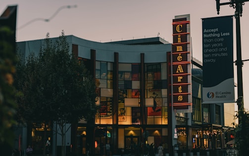 A theater building with a prominent vertical neon sign displaying 'Ciné Arts' situated at the front. The structure features large glass windows and is surrounded by trees and street lamps. A banner for El Camino Health hangs nearby with a motivational message.
