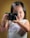 A young child with long hair is holding a camera and looking through the viewfinder. The child is wearing a white top with the word 'California' printed in purple. A small purple hair clip holds part of the hair in place. The background is a solid, muted yellowish-brown color.