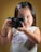 A young child with long hair is holding a camera and looking through the viewfinder. The child is wearing a white top with the word 'California' printed in purple. A small purple hair clip holds part of the hair in place. The background is a solid, muted yellowish-brown color.