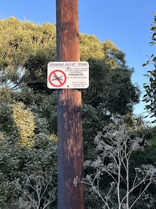 A sign posted on a wooden pole amidst greenery warns against the use of unmanned aircraft drones. The sign contains a red circle with a diagonal line over a drone icon, symbolizing prohibition.