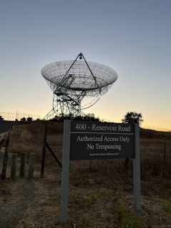 A large satellite dish set against a twilight sky with a sign in the foreground reading '400 - Reservoir Road Authorized Access Only No Trespassing.' The ground is covered in dry grass, and there is a fence surrounding the area.