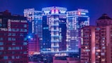 A cityscape at night featuring tall buildings illuminated with neon lights in various shades of blue and pink. The buildings have multiple signs and words displayed across them, creating a futuristic and modern aesthetic. The sky is a deep purple, adding to the cyberpunk atmosphere.