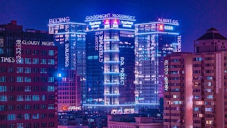 A cityscape at night featuring tall buildings illuminated with neon lights in various shades of blue and pink. The buildings have multiple signs and words displayed across them, creating a futuristic and modern aesthetic. The sky is a deep purple, adding to the cyberpunk atmosphere.