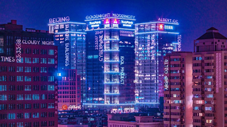 A cityscape at night featuring tall buildings illuminated with neon lights in various shades of blue and pink. The buildings have multiple signs and words displayed across them, creating a futuristic and modern aesthetic. The sky is a deep purple, adding to the cyberpunk atmosphere.