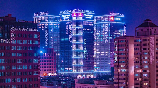A cityscape at night featuring tall buildings illuminated with neon lights in various shades of blue and pink. The buildings have multiple signs and words displayed across them, creating a futuristic and modern aesthetic. The sky is a deep purple, adding to the cyberpunk atmosphere.