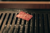 A piece of marbled meat is placed on a black grill with visible grill lines. The surface of the grill is shiny, likely due to grease or oil.