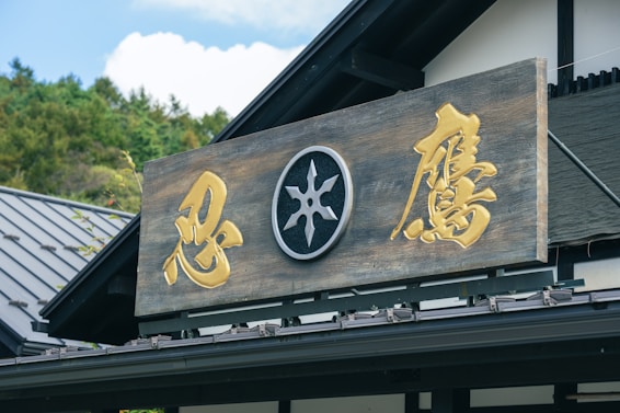 A wooden sign with gold-colored Japanese characters and a central emblem, mounted on the exterior of a traditional-style building. The roof is visible, with a sloped modern design, and greenery can be seen in the background under a clear sky.