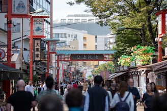A bustling street scene filled with pedestrians walking under a series of decorative red arches. Traditional Japanese architecture is visible on either side of the street, and the area is lined with shops and vendors. The atmosphere is lively with a mix of both locals and tourists.