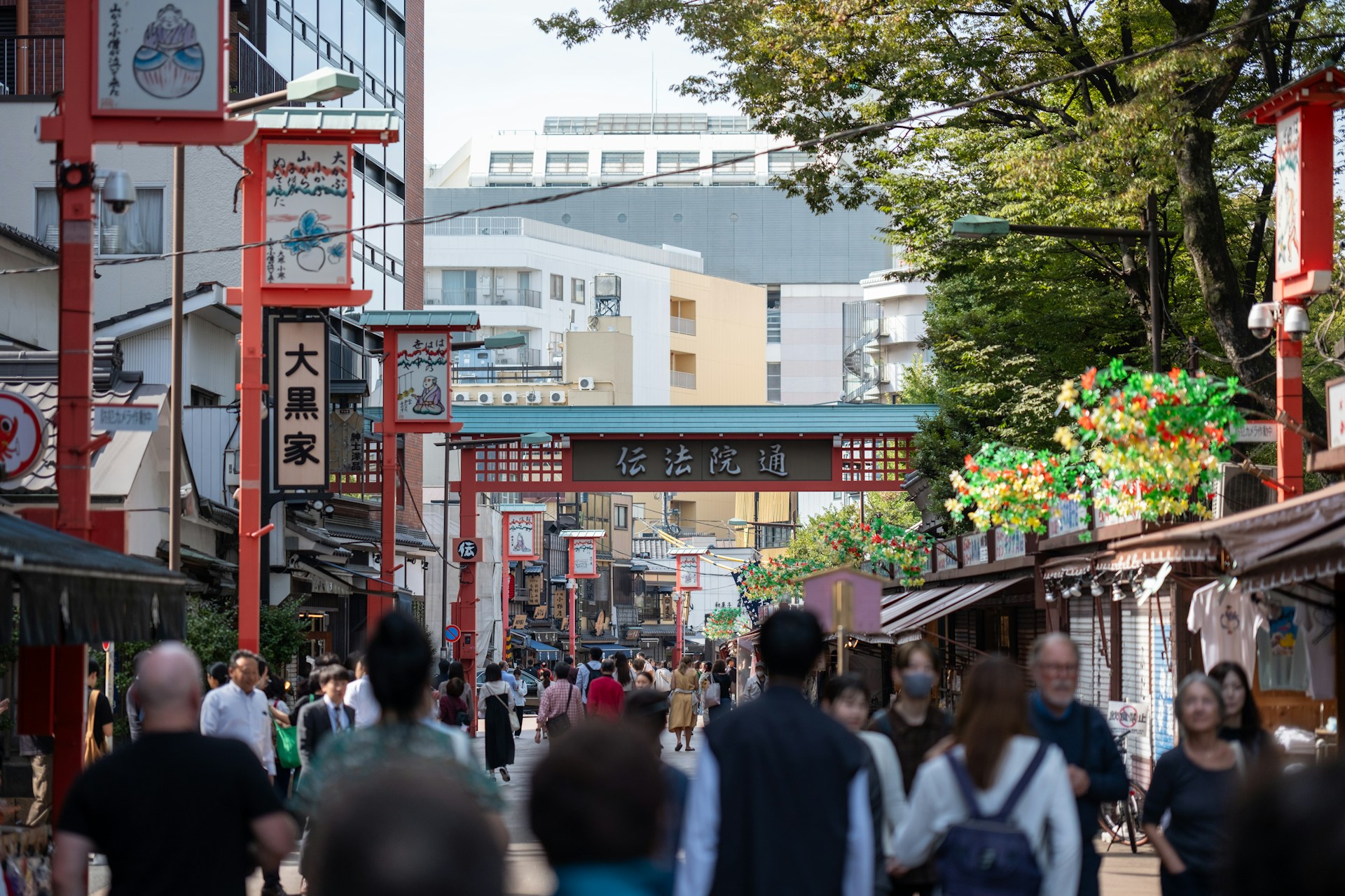 A bustling street scene filled with pedestrians walking under a series of decorative red arches. Traditional Japanese architecture is visible on either side of the street, and the area is lined with shops and vendors. The atmosphere is lively with a mix of both locals and tourists.