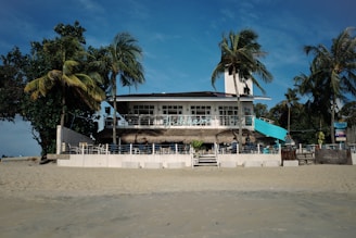 A beach house with a light blue and white facade is surrounded by palm trees and directly faces the sandy shore. The house features a second-floor terrace with an outdoor seating area and a few people are visible enjoying the space under a bright blue sky.