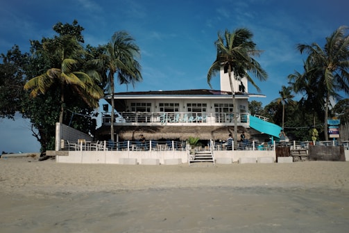 A beach house with a light blue and white facade is surrounded by palm trees and directly faces the sandy shore. The house features a second-floor terrace with an outdoor seating area and a few people are visible enjoying the space under a bright blue sky.