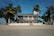 A beach house with a light blue and white facade is surrounded by palm trees and directly faces the sandy shore. The house features a second-floor terrace with an outdoor seating area and a few people are visible enjoying the space under a bright blue sky.