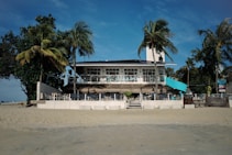 A beach house with a light blue and white facade is surrounded by palm trees and directly faces the sandy shore. The house features a second-floor terrace with an outdoor seating area and a few people are visible enjoying the space under a bright blue sky.