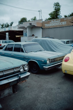 A row of vintage cars is parked outdoors near a repair shop, with one car covered by a tarp. The repair shop has a sign that reads 'Inland Transmissions'. The vehicles are mostly in shades of blue and yellow. It's a cloudy day, with trees visible in the background.
