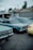 A row of vintage cars is parked outdoors near a repair shop, with one car covered by a tarp. The repair shop has a sign that reads 'Inland Transmissions'. The vehicles are mostly in shades of blue and yellow. It's a cloudy day, with trees visible in the background.