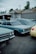 A row of vintage cars is parked outdoors near a repair shop, with one car covered by a tarp. The repair shop has a sign that reads 'Inland Transmissions'. The vehicles are mostly in shades of blue and yellow. It's a cloudy day, with trees visible in the background.