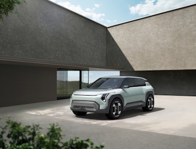 A modern, sleek SUV is parked on a concrete driveway beside a minimalist building. The vehicle features a futuristic design with angular lines and large wheels. The building has textured walls and expansive windows that reflect the surrounding environment. Greenery is visible in the foreground, suggesting a blend of nature and urban design.
