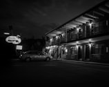 A vintage-style motel with a large sign reading 'Apache Motel' is illuminated at night. The two-story brick building features outdoor lighting and a row of windows and doors. A parked car is visible in the foreground, and the scene is captured in black and white, adding a nostalgic atmosphere.