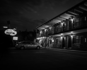 A vintage-style motel with a large sign reading 'Apache Motel' is illuminated at night. The two-story brick building features outdoor lighting and a row of windows and doors. A parked car is visible in the foreground, and the scene is captured in black and white, adding a nostalgic atmosphere.