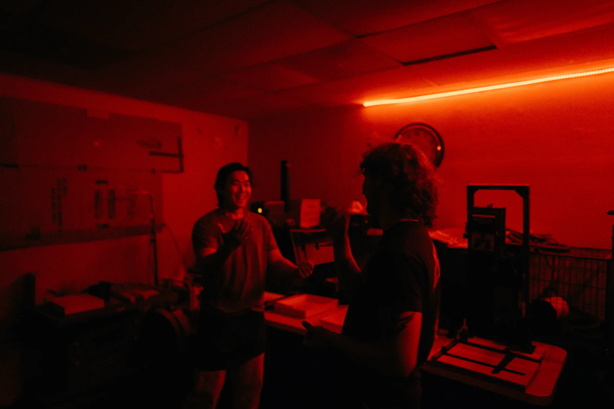 Deux jeunes hommes rient face à face dans une pièce plongée dans une lumière rouge, une horloge ronde accrochée au mur