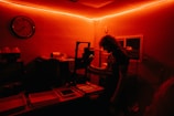 A person stands inside a dimly lit darkroom illuminated with red light. The individual appears to be working with photographic development equipment, including trays and an enlarger. A wall clock and bottles with chemicals are visible in the background.