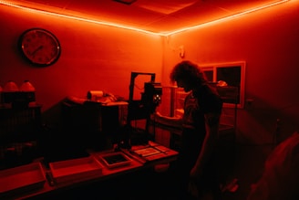 A person stands inside a dimly lit darkroom illuminated with red light. The individual appears to be working with photographic development equipment, including trays and an enlarger. A wall clock and bottles with chemicals are visible in the background.