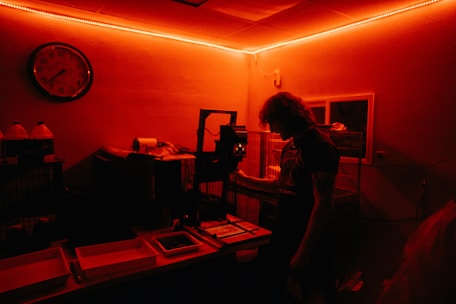 A person stands inside a dimly lit darkroom illuminated with red light. The individual appears to be working with photographic development equipment, including trays and an enlarger. A wall clock and bottles with chemicals are visible in the background.