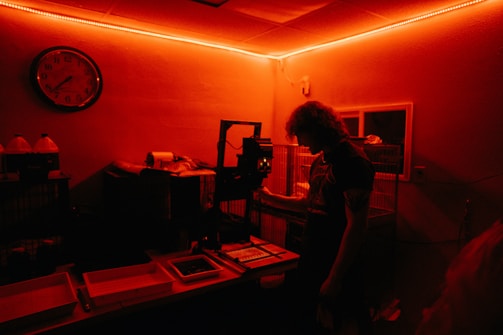 A person stands inside a dimly lit darkroom illuminated with red light. The individual appears to be working with photographic development equipment, including trays and an enlarger. A wall clock and bottles with chemicals are visible in the background.