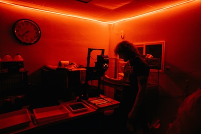 A person stands inside a dimly lit darkroom illuminated with red light. The individual appears to be working with photographic development equipment, including trays and an enlarger. A wall clock and bottles with chemicals are visible in the background.