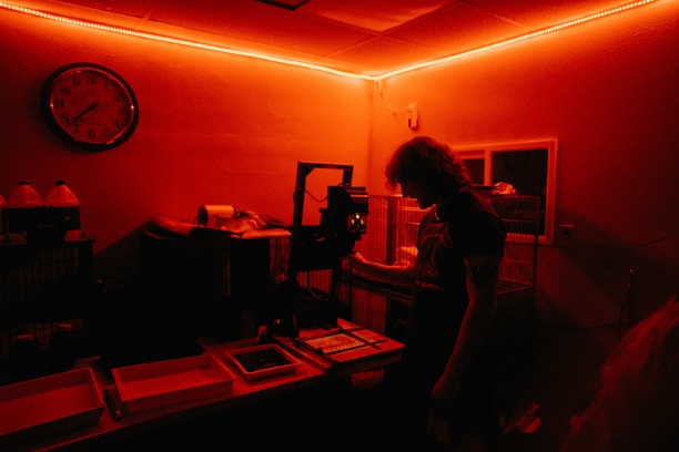A person stands inside a dimly lit darkroom illuminated with red light. The individual appears to be working with photographic development equipment, including trays and an enlarger. A wall clock and bottles with chemicals are visible in the background.