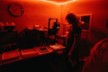 A dimly lit room with a red hue. A person with curly hair stands in front of a table, possibly in a darkroom environment for developing photographs. Various trays, bottles, and equipment are visible on the table. The room has a vintage clock on the wall and cages are lined up against the wall.