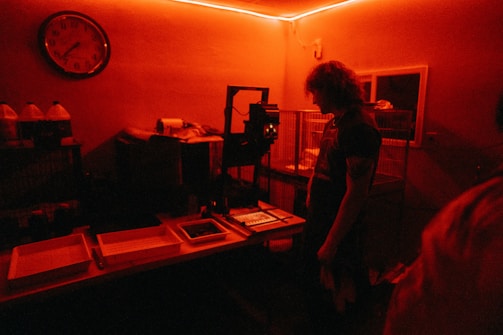 A dimly lit room with a red hue. A person with curly hair stands in front of a table, possibly in a darkroom environment for developing photographs. Various trays, bottles, and equipment are visible on the table. The room has a vintage clock on the wall and cages are lined up against the wall.