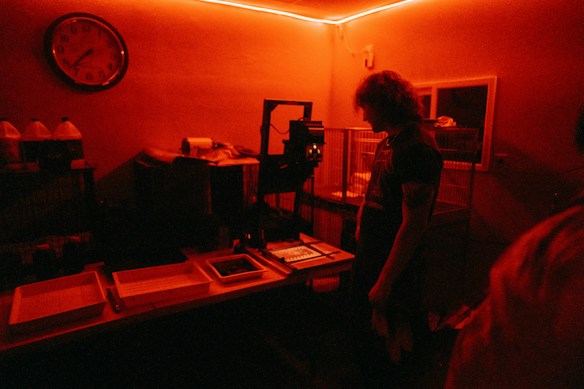 A dimly lit room with a red hue. A person with curly hair stands in front of a table, possibly in a darkroom environment for developing photographs. Various trays, bottles, and equipment are visible on the table. The room has a vintage clock on the wall and cages are lined up against the wall.