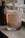 A lit scented candle in a glass jar is placed on a surface near a window with blinds. The candle has a label that reads 'Stone & Ivy Sweater Weather' with scents listed as blood orange, cinnamon, and rose. Soft ambient lighting creates a warm and cozy atmosphere.