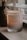 A lit scented candle in a glass jar is placed on a surface near a window with blinds. The candle has a label that reads 'Stone & Ivy Sweater Weather' with scents listed as blood orange, cinnamon, and rose. Soft ambient lighting creates a warm and cozy atmosphere.