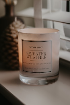 A lit scented candle in a glass jar is placed on a surface near a window with blinds. The candle has a label that reads 'Stone & Ivy Sweater Weather' with scents listed as blood orange, cinnamon, and rose. Soft ambient lighting creates a warm and cozy atmosphere.