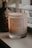 A lit scented candle in a glass jar is placed on a surface near a window with blinds. The candle has a label that reads 'Stone & Ivy Sweater Weather' with scents listed as blood orange, cinnamon, and rose. Soft ambient lighting creates a warm and cozy atmosphere.
