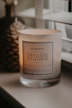 A lit scented candle in a glass jar is placed on a surface near a window with blinds. The candle has a label that reads 'Stone & Ivy Sweater Weather' with scents listed as blood orange, cinnamon, and rose. Soft ambient lighting creates a warm and cozy atmosphere.
