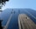 Several workers in harnesses are suspended on ropes while cleaning the glass facade of a skyscraper. The building reflects another skyscraper and the clear blue sky. A tree with green leaves is partially visible in the upper left corner.