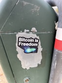A sticker is placed on a green metal surface next to a metal pole. The sticker features the phrase 'Bitcoin is Freedom' with a graphic of abstract shapes, including a blue-green swoosh and small stars.