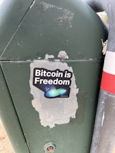 A sticker is placed on a green metal surface next to a metal pole. The sticker features the phrase 'Bitcoin is Freedom' with a graphic of abstract shapes, including a blue-green swoosh and small stars.