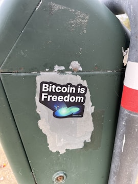 A sticker is placed on a green metal surface next to a metal pole. The sticker features the phrase 'Bitcoin is Freedom' with a graphic of abstract shapes, including a blue-green swoosh and small stars.